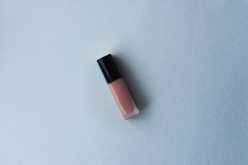 A tube of lip gloss with a black cap and pink hue is placed on a light blue textured surface. The minimalist composition creates a simple and clean aesthetic.