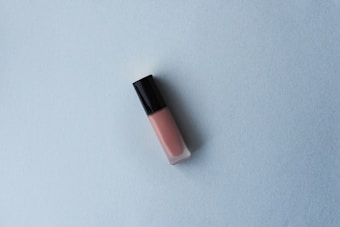 A tube of lip gloss with a black cap and pink hue is placed on a light blue textured surface. The minimalist composition creates a simple and clean aesthetic.