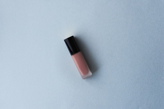 A tube of lip gloss with a black cap and pink hue is placed on a light blue textured surface. The minimalist composition creates a simple and clean aesthetic.
