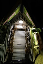 A closed florist shop with a metallic roll-up shutter surrounded by decorative dried branches. The storefront has a distinct black and white striped design with green neon signs. The scene is captured at night, and a bright round light fixture illuminates the entrance.