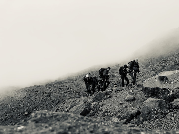 A group of hikers with backpacks is traversing a rocky, foggy mountain slope. The terrain is rugged and uneven, filled with rocks and pebbles, while a thick mist envelops the background, creating a sense of isolation and adventure.