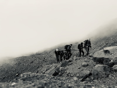 A group of hikers with backpacks is traversing a rocky, foggy mountain slope. The terrain is rugged and uneven, filled with rocks and pebbles, while a thick mist envelops the background, creating a sense of isolation and adventure.