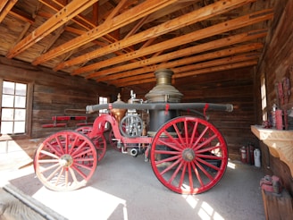 An old-fashioned fire engine with large red wheels and a brass water tank is stored in a wooden shed with exposed beams. The bright sunlight illuminates the scene through windows, casting shadows on the ground. Nearby are firefighting tools and equipment including extinguishers and other historical items.