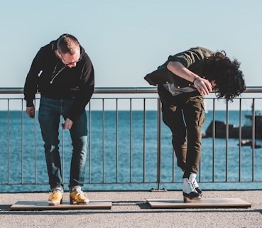 Two individuals are engaged in a tap dance routine on a promenade by the sea. They are leaning forward with energy, each positioned on a small wooden board. The background features a clear blue sky and a deep blue ocean, with a metal railing separating the dancers from the water.