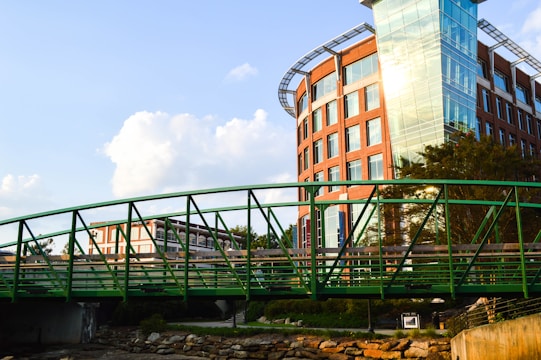 A modern building with a glass facade and orange brickwork stands adjacent to a green metal pedestrian bridge. The building has a rounded corner and multiple windows. Trees and neatly arranged landscaping surround the structure. The sky is clear with a few fluffy clouds, indicating good weather.