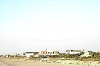 Several houses line a sandy beach, with wooden fences and greenery in the foreground. The sky is clear and light in color, giving the scene a serene coastal vibe.