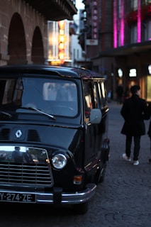A vintage black van is parked on a cobblestone street in an urban setting. Bright neon signs light up the background, and two people walk closely together, creating a lively and urban nightlife atmosphere.