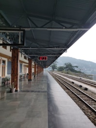A railway platform with a digital clock displaying 12:37. The platform is lined with potted plants and has a long roof supported by wooden pillars. Train tracks run alongside the platform with distant hills visible under an overcast sky.