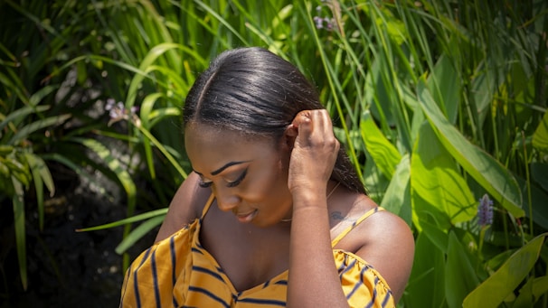 A person with smooth, dark hair is gently touching their ear while looking down, set against a backdrop of lush green foliage and intermittent flowers. They are wearing a yellow striped top, revealing an off-the-shoulder style.