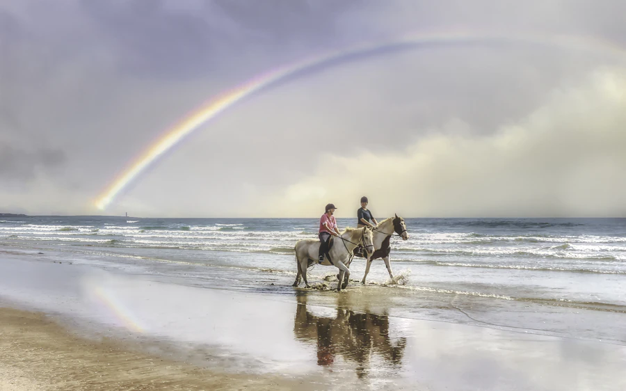 Two riders on horseback along a beach with a rainbow