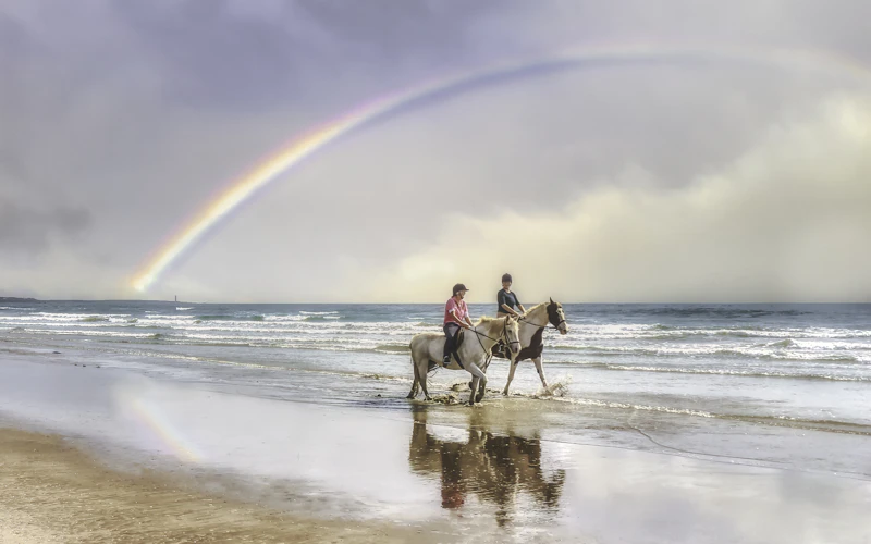 Two riders on horseback along the shoreline beneath a rainbow