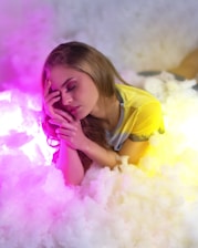 A woman with long hair rests peacefully among soft, cloud-like surroundings, with vibrant pink and yellow hues casting a dreamy light over the scene. Her eyes are closed, and her hand gently touches her face, creating a serene and tranquil atmosphere.