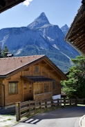 A picturesque wooden chalet sits in a serene mountain setting with a towering, rocky peak in the background. The house has a sloped, shingled roof and multiple windows, surrounded by lush greenery and a clear blue sky.