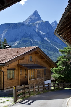 A picturesque wooden chalet sits in a serene mountain setting with a towering, rocky peak in the background. The house has a sloped, shingled roof and multiple windows, surrounded by lush greenery and a clear blue sky.