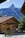 A picturesque wooden chalet sits in a serene mountain setting with a towering, rocky peak in the background. The house has a sloped, shingled roof and multiple windows, surrounded by lush greenery and a clear blue sky.