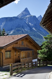 A picturesque wooden chalet sits in a serene mountain setting with a towering, rocky peak in the background. The house has a sloped, shingled roof and multiple windows, surrounded by lush greenery and a clear blue sky.