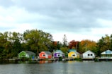 A row of colorful cottages lines the edge of a calm body of water, set against a backdrop of trees in autumn foliage. Overcast skies cast a muted light over the vibrant scene, with each cottage painted a distinct color including green, red, purple, yellow, and white.