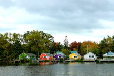 A row of colorful cottages lines the edge of a calm body of water, set against a backdrop of trees in autumn foliage. Overcast skies cast a muted light over the vibrant scene, with each cottage painted a distinct color including green, red, purple, yellow, and white.