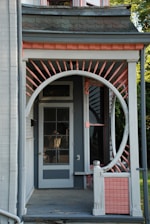 A porch features a unique circular entrance frame surrounded by decorative pink and white spindles, leading up to a glass-paneled door. The porch is part of a house with distinctive architectural elements, including shingles and a light grey color scheme accented by pink. Parts of the roofline and siding are visible, along with some greenery in the background.