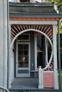 A porch features a unique circular entrance frame surrounded by decorative pink and white spindles, leading up to a glass-paneled door. The porch is part of a house with distinctive architectural elements, including shingles and a light grey color scheme accented by pink. Parts of the roofline and siding are visible, along with some greenery in the background.