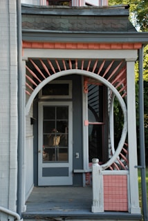 A porch features a unique circular entrance frame surrounded by decorative pink and white spindles, leading up to a glass-paneled door. The porch is part of a house with distinctive architectural elements, including shingles and a light grey color scheme accented by pink. Parts of the roofline and siding are visible, along with some greenery in the background.