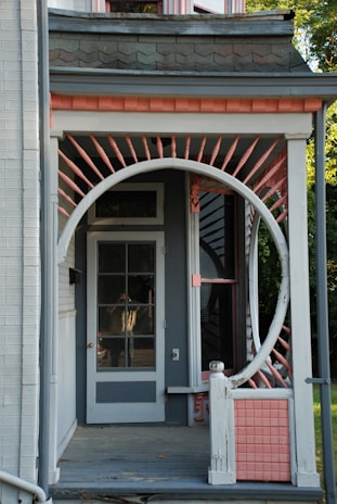 A porch features a unique circular entrance frame surrounded by decorative pink and white spindles, leading up to a glass-paneled door. The porch is part of a house with distinctive architectural elements, including shingles and a light grey color scheme accented by pink. Parts of the roofline and siding are visible, along with some greenery in the background.