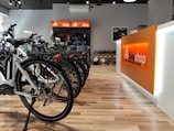 A modern bike shop interior with several bicycles lined up on a wooden floor. The shop has a bright and clean aesthetic, with an orange and white color scheme. Bike accessories and helmets are visible on display shelves behind the counter.