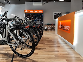 A modern bike shop interior with several bicycles lined up on a wooden floor. The shop has a bright and clean aesthetic, with an orange and white color scheme. Bike accessories and helmets are visible on display shelves behind the counter.