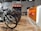 A modern bike shop interior with several bicycles lined up on a wooden floor. The shop has a bright and clean aesthetic, with an orange and white color scheme. Bike accessories and helmets are visible on display shelves behind the counter.