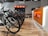 A modern bike shop interior with several bicycles lined up on a wooden floor. The shop has a bright and clean aesthetic, with an orange and white color scheme. Bike accessories and helmets are visible on display shelves behind the counter.