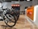 A modern bike shop interior with several bicycles lined up on a wooden floor. The shop has a bright and clean aesthetic, with an orange and white color scheme. Bike accessories and helmets are visible on display shelves behind the counter.