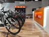 A modern bike shop interior with several bicycles lined up on a wooden floor. The shop has a bright and clean aesthetic, with an orange and white color scheme. Bike accessories and helmets are visible on display shelves behind the counter.