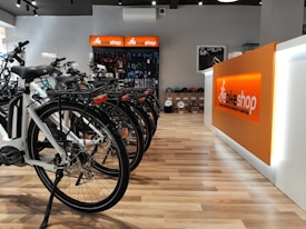 A modern bike shop interior with several bicycles lined up on a wooden floor. The shop has a bright and clean aesthetic, with an orange and white color scheme. Bike accessories and helmets are visible on display shelves behind the counter.