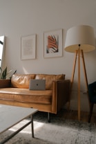A cozy living room features a tan leather couch with a laptop resting on it. Above the couch, two framed abstract art prints hang on the wall. A tall floor lamp with a beige lampshade and wooden tripod base stands to the right of the couch. Natural light flows in from a window on the left, creating a serene ambiance.
