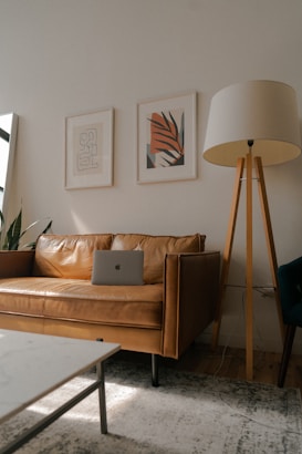 A cozy living room features a tan leather couch with a laptop resting on it. Above the couch, two framed abstract art prints hang on the wall. A tall floor lamp with a beige lampshade and wooden tripod base stands to the right of the couch. Natural light flows in from a window on the left, creating a serene ambiance.