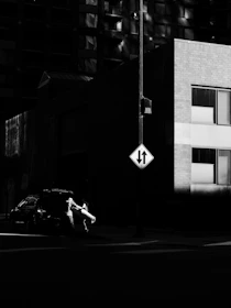A black and white image of a person loading or unloading a large item from a car parked on a city street. The scene is dominated by shadows, with a bright light illuminating the person and the object they are handling. A building and a traffic sign are visible in the background, with the sign indicating traffic directions.