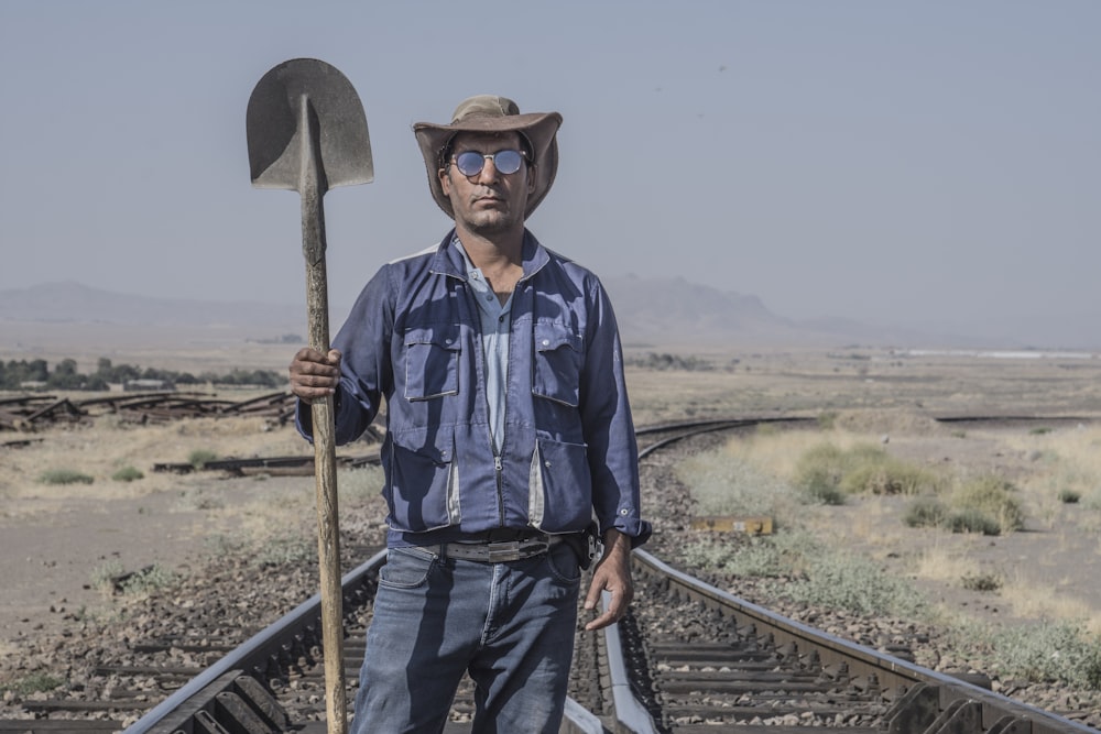 homem na camisa social xadrez azul e branca e chapéu de cowboy marrom segurando pá de madeira marrom