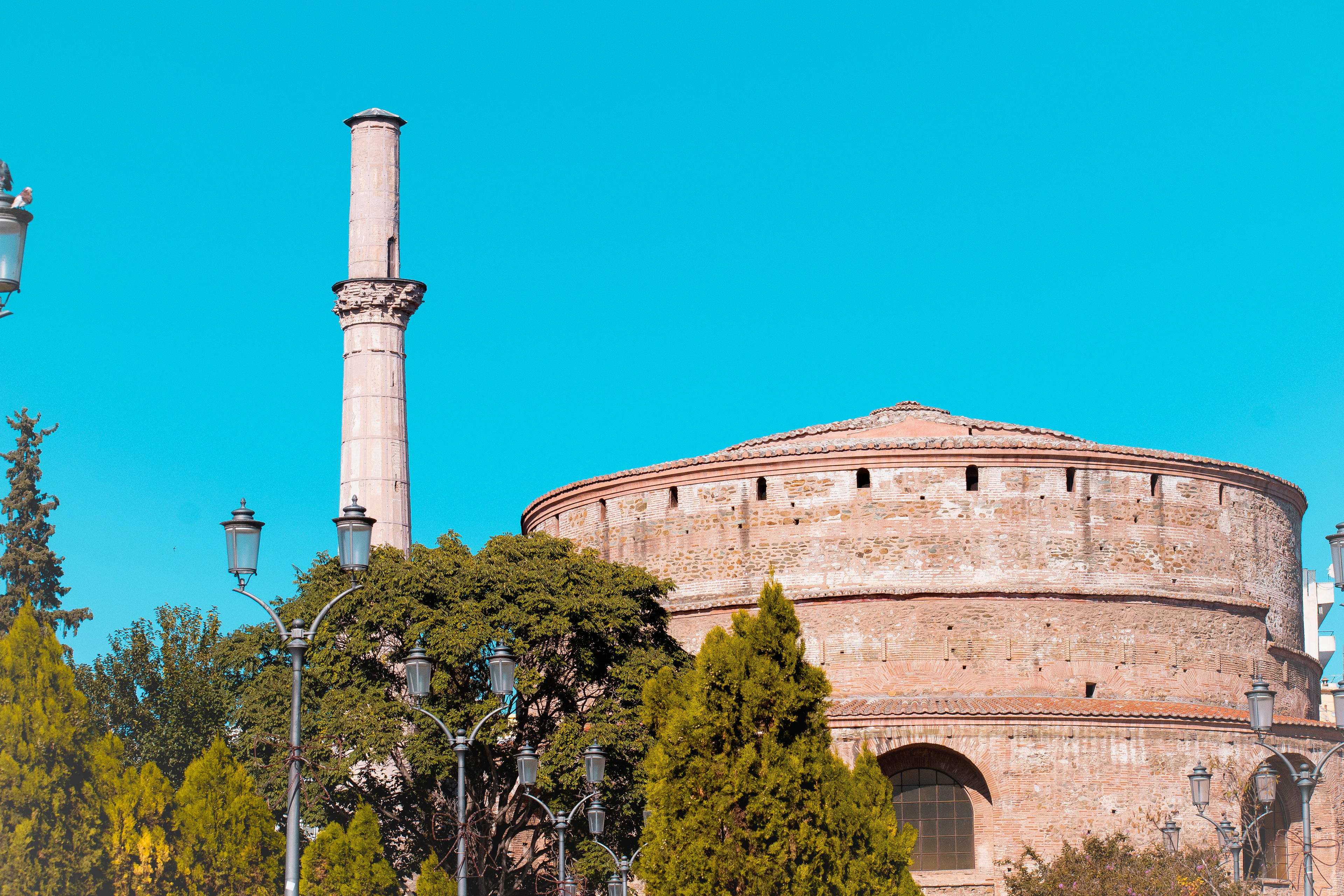 The vast brick drum of the Rotunda of Galerius in Thessaloniki beside its surviving Ottoman minaret, cypresses and street lamps below a deep blue sky.