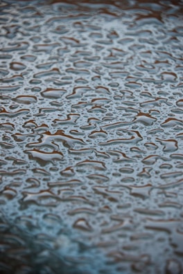 Surface covered with numerous water droplets forming irregular patterns. The droplets are clear and reflective, creating a textured appearance against a slightly blurred background.