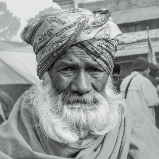 An elderly man with a long white beard, wearing a turban with patterns and text, looks directly at the viewer. His expression is serious and contemplative, with deep lines on his face reflecting age and wisdom. In the background, other people are blurred, wearing winter clothing, suggesting a cold setting.