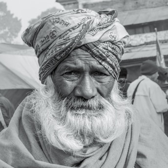 An elderly man with a long white beard, wearing a turban with patterns and text, looks directly at the viewer. His expression is serious and contemplative, with deep lines on his face reflecting age and wisdom. In the background, other people are blurred, wearing winter clothing, suggesting a cold setting.