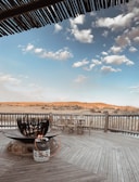 A wooden deck features a large circular fire pit with a gas tank connected via a hose. There are rustic wooden chairs arranged near the railing, which overlooks an expansive landscape of sandy terrain and sparse vegetation. The sky above is filled with light, fluffy clouds against a clear blue backdrop. Wooden roofing provides partial shade in the foreground.