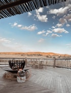 A wooden deck features a large circular fire pit with a gas tank connected via a hose. There are rustic wooden chairs arranged near the railing, which overlooks an expansive landscape of sandy terrain and sparse vegetation. The sky above is filled with light, fluffy clouds against a clear blue backdrop. Wooden roofing provides partial shade in the foreground.
