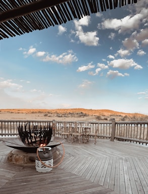 A wooden deck features a large circular fire pit with a gas tank connected via a hose. There are rustic wooden chairs arranged near the railing, which overlooks an expansive landscape of sandy terrain and sparse vegetation. The sky above is filled with light, fluffy clouds against a clear blue backdrop. Wooden roofing provides partial shade in the foreground.