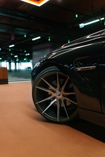 A close-up view of a car's wheel in a dimly lit parking garage. The focus is on the sleek, metallic alloy wheel of the vehicle, with the background including ceiling lights and other parked cars. The garage floor has a smooth, tan color.