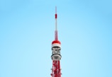 A tall, red and white communications tower against a clear blue sky. The tower features intricate metalwork and multiple antennae, demonstrating its role as a broadcasting or observation tower.