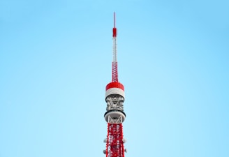 A tall, red and white communications tower against a clear blue sky. The tower features intricate metalwork and multiple antennae, demonstrating its role as a broadcasting or observation tower.