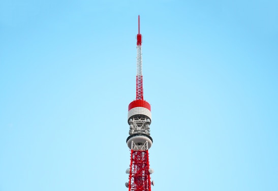 A tall, red and white communications tower against a clear blue sky. The tower features intricate metalwork and multiple antennae, demonstrating its role as a broadcasting or observation tower.