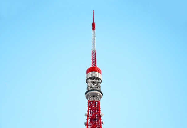 A tall, red and white communications tower against a clear blue sky. The tower features intricate metalwork and multiple antennae, demonstrating its role as a broadcasting or observation tower.