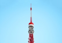 A tall, red and white communications tower against a clear blue sky. The tower features intricate metalwork and multiple antennae, demonstrating its role as a broadcasting or observation tower.